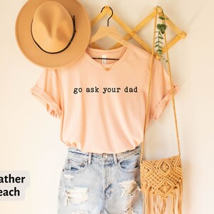 May include: A peach coloured t-shirt with the text "go ask your dad" in black lettering. The shirt is hanging on a wooden hanger and has a brown hat and a woven bag next to it.