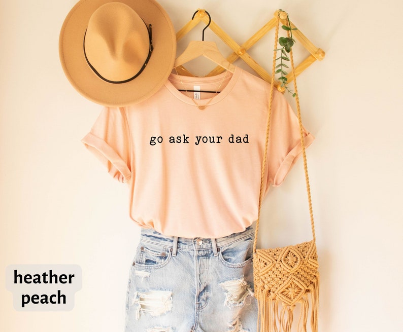 May include: A peach coloured t-shirt with the text "go ask your dad" in black lettering. The shirt is hanging on a wooden hanger and has a brown hat and a woven bag next to it.