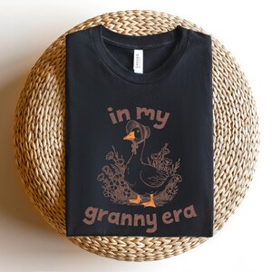May include: Black t-shirt with the text "in my granny era" and a graphic of a goose wearing a bonnet. The goose is surrounded by flowers. The shirt is displayed on a round, woven surface.