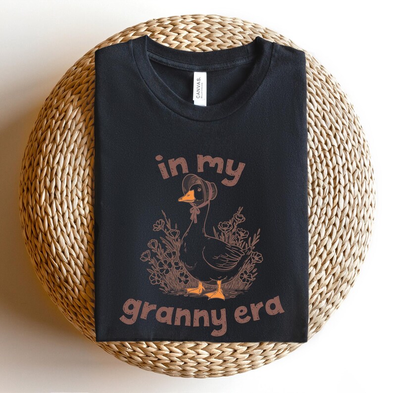 May include: Black t-shirt with the text "in my granny era" and a graphic of a goose wearing a bonnet. The goose is surrounded by flowers. The shirt is displayed on a round, woven surface.