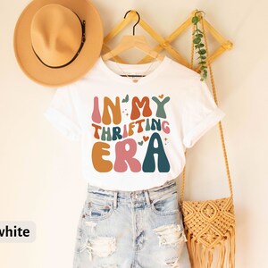May include: White t-shirt with the text "IN MY THRIFTING ERA" in retro-style lettering. The design features various colors, including orange, pink, teal, and brown. The shirt is hanging on a wooden hanger, paired with denim shorts and a tan hat.