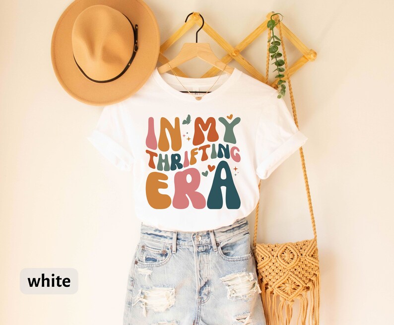 May include: White t-shirt with the text "IN MY THRIFTING ERA" in retro-style lettering. The design features various colors, including orange, pink, teal, and brown. The shirt is hanging on a wooden hanger, paired with denim shorts and a tan hat.