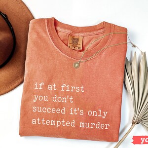 Op de afbeelding: Een koraalkleurig Comfort Colors T-shirt met de tekst "if at first you don't succeed it's only attempted murder" in witte letters. Een gouden ketting en een bruine hoed liggen naast het shirt.