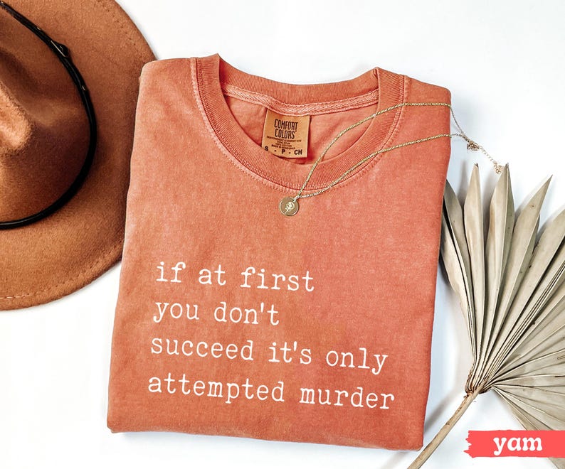 Op de afbeelding: Een koraalkleurig Comfort Colors T-shirt met de tekst "if at first you don't succeed it's only attempted murder" in witte letters. Een gouden ketting en een bruine hoed liggen naast het shirt.