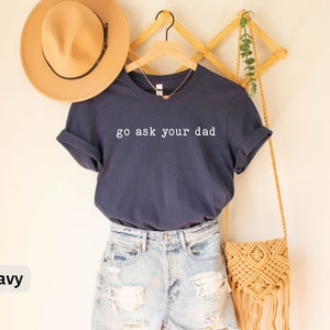 May include: Navy blue t-shirt with the text "go ask your dad" in white. The shirt is hanging on a wooden rack with a straw bag and a hat.