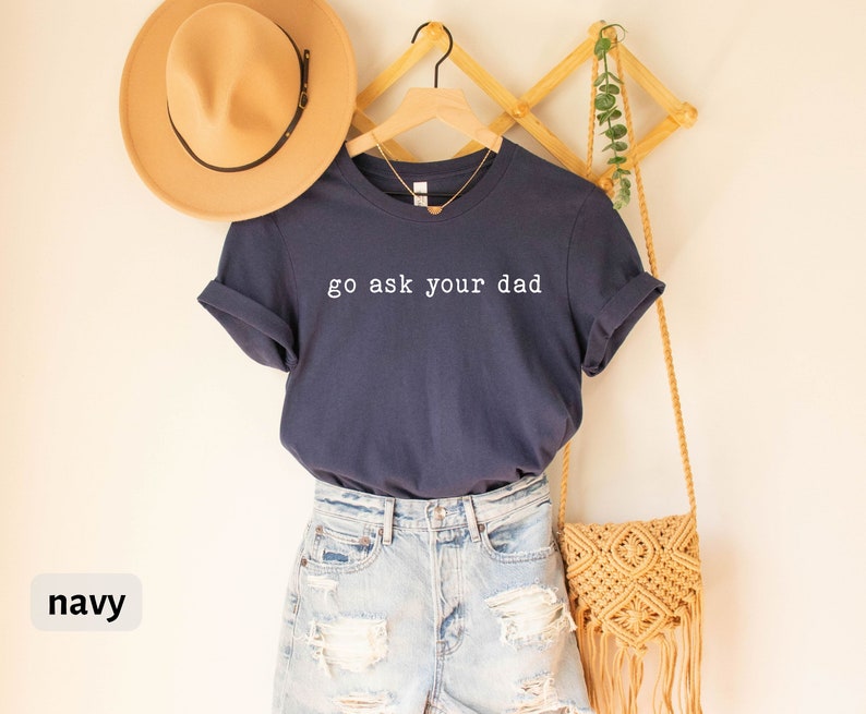 May include: Navy blue t-shirt with the text "go ask your dad" in white. The shirt is hanging on a wooden rack with a straw bag and a hat.