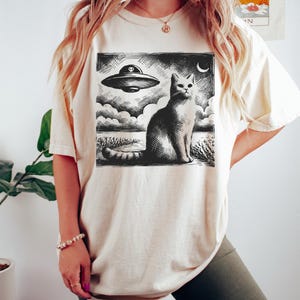 May include: A white oversized t-shirt with a black and white graphic print of a cat sitting in front of a flying saucer. The graphic is in a vintage woodcut style.