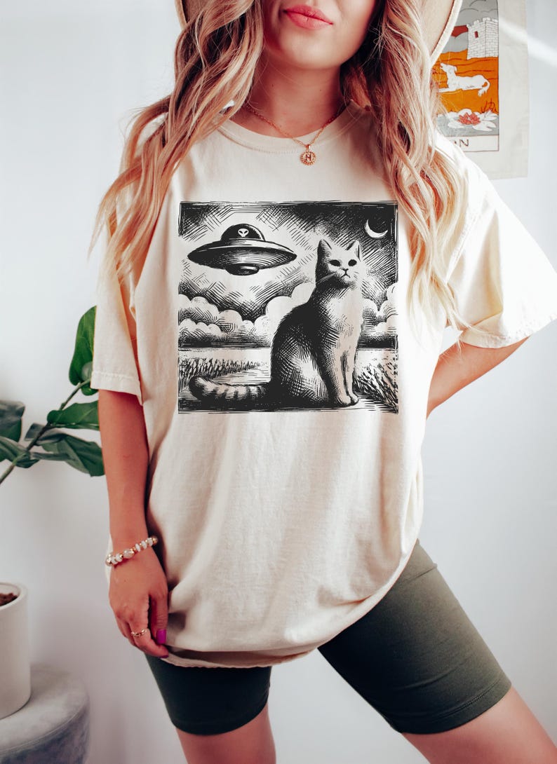 May include: A white oversized t-shirt with a black and white graphic print of a cat sitting in front of a flying saucer. The graphic is in a vintage woodcut style.