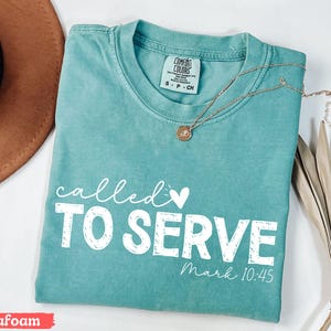 Pu&ograve; includere: T-shirt verde acqua con le parole "called TO SERVE" in bianco, insieme a "Mark 10:45". Una collana d'oro con un ciondolo &egrave; appoggiata sulla maglietta. La maglietta &egrave; piegata e ha un'etichetta Comfort Colors.