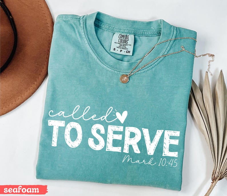 Pu&ograve; includere: T-shirt verde acqua con le parole "called TO SERVE" in bianco, insieme a "Mark 10:45". Una collana d'oro con un ciondolo &egrave; appoggiata sulla maglietta. La maglietta &egrave; piegata e ha un'etichetta Comfort Colors.