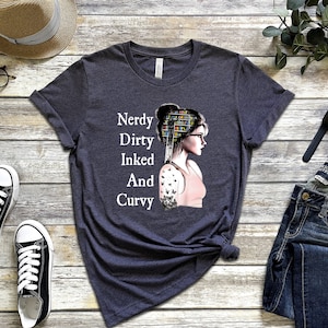 Op de afbeelding: Een donker grijs t-shirt met een afbeelding van een vrouw met boeken op haar hoofd en een vlinder-tatoeage op haar arm. De tekst "Nerdy Dirty Inked And Curvy" staat op het shirt gedrukt.