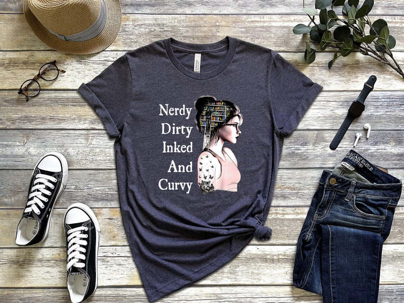 Op de afbeelding: Een donker grijs t-shirt met een afbeelding van een vrouw met boeken op haar hoofd en een vlinder-tatoeage op haar arm. De tekst "Nerdy Dirty Inked And Curvy" staat op het shirt gedrukt.