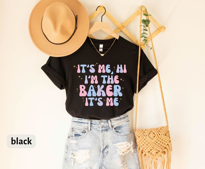 Peut inclure: T-shirt noir avec le texte "It's me, hi I'm the baker It's me" en lettres roses et bleues avec des accents de paillettes. Le t-shirt est suspendu &agrave; un support en bois avec un sac en paille et une plante.