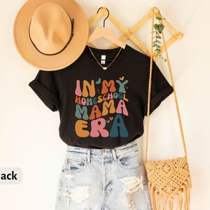 May include: Black t-shirt with the text "In My Homeschool Mama Era" in colorful, retro-style lettering. The shirt is hanging on a wooden rack with a straw hat and a woven bag.
