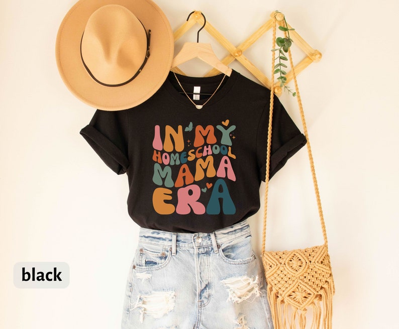 May include: Black t-shirt with the text "In My Homeschool Mama Era" in colorful, retro-style lettering. The shirt is hanging on a wooden rack with a straw hat and a woven bag.