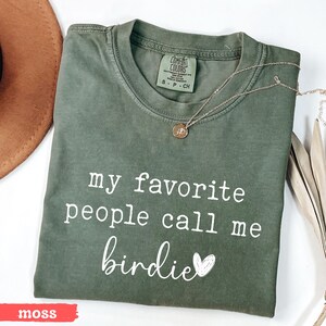May include: A moss green t-shirt with the text "my favourite people call me birdie" in white script. A gold necklace with a pendant is on the shirt. The shirt is folded and next to a brown hat.