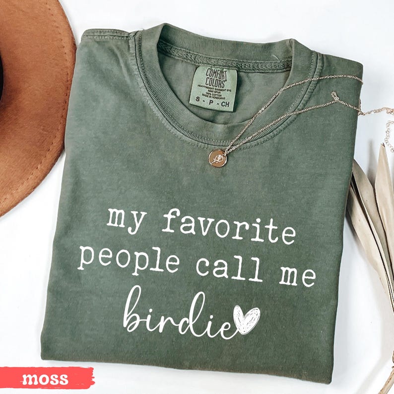 May include: A moss green t-shirt with the text "my favourite people call me birdie" in white script. A gold necklace with a pendant is on the shirt. The shirt is folded and next to a brown hat.
