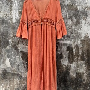 May include: A long-sleeved, burnt orange duster cardigan featuring a deep V-neckline, flared bell sleeves, and a wide, intricate crochet-style lace panel across the waist. The garment includes two small buttons at the centre front closure.