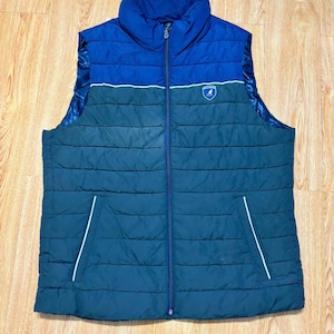 Op de afbeelding: Een mouwloos gewatteerd vest met een tweekleurig ontwerp, bestaande uit een felblauw bovendeel en een donkergroenblauw onderste deel. Het vest is voorzien van een ritssluiting over de gehele lengte, een hoge kraag en een kangoeroe-logo op de borst.