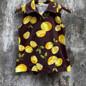May include: A sleeveless collared shirt in a dark brown color featuring a repeating pattern of bright yellow lemons and green leaves. The shirt has a pointed collar, a buttonless placket, and a straight hemline, showcasing a vibrant fruit print.