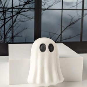 May include: A white ghost-shaped bath bomb with two black eyes. The bath bomb is sitting on a white rectangular block. The background features a window with a spooky night scene.