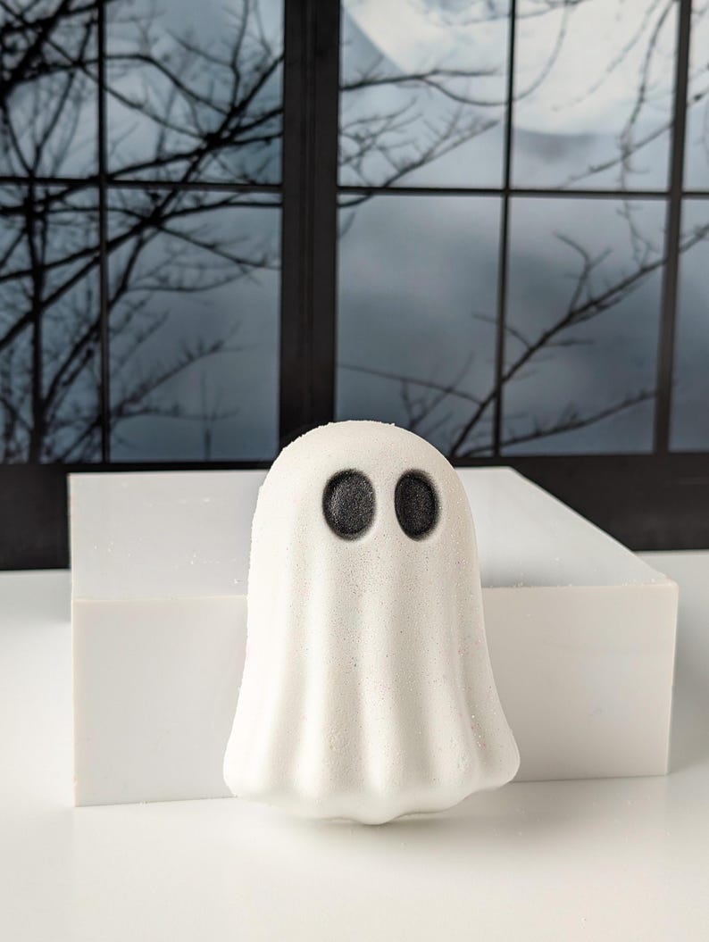 May include: A white ghost-shaped bath bomb with two black eyes. The bath bomb is sitting on a white rectangular block. The background features a window with a spooky night scene.