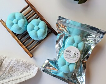 May include: Two blue shower steamers shaped like flowers, one in a wooden rack and one in a silver pouch. The pouch has a label that reads "Breathe Peppermint & Eucalyptus Shower Steamers".