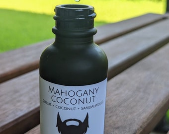 May include: A black bottle of beard oil with a white label that says "Mahogany Coconut" and "Beard Oil". The label also lists the ingredients: "Citrus + Coconut + Sandalwood". The bottle is 1 ounce and contains 32 grams of oil.