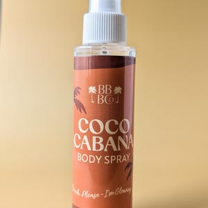 Puede incluir: Un frasco transparente de spray corporal con un pulverizador blanco. La etiqueta es marr&oacute;n con las palabras "COCO CABANA BODY SPRAY" en blanco. La botella contiene 118 ml de producto. El texto "Beach, Please - I'm Glowing" tambi&eacute;n est&aacute; en la etiqueta.