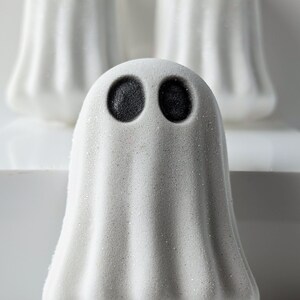 May include: Three white ghost-shaped bath bombs with black eyes are arranged on a white surface. The front-most ghost is in focus, with the other two slightly blurred in the background. The bath bombs have a textured, glittery appearance, perfect for Halloween.