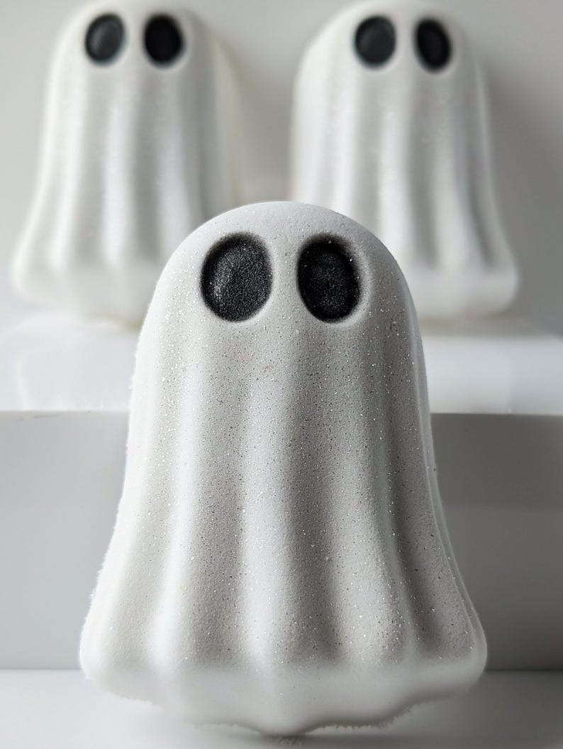May include: Three white ghost-shaped bath bombs with black eyes are arranged on a white surface. The front-most ghost is in focus, with the other two slightly blurred in the background. The bath bombs have a textured, glittery appearance, perfect for Halloween.