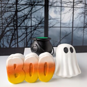 May include: A Halloween-themed arrangement featuring a black cauldron, the word "BOO" in candy corn colors, and a white ghost figure with black eyes. The items are arranged on a white surface against a backdrop of a window with a spooky scene.