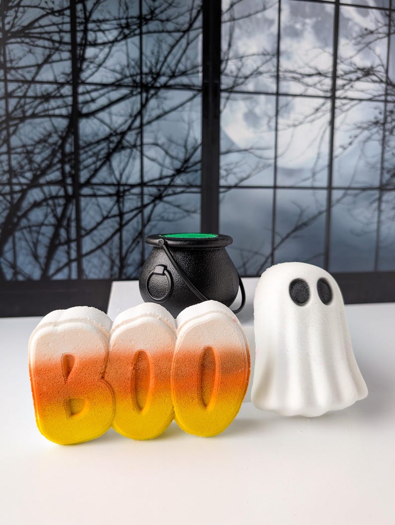 May include: A Halloween-themed arrangement featuring a black cauldron, the word "BOO" in candy corn colors, and a white ghost figure with black eyes. The items are arranged on a white surface against a backdrop of a window with a spooky scene.