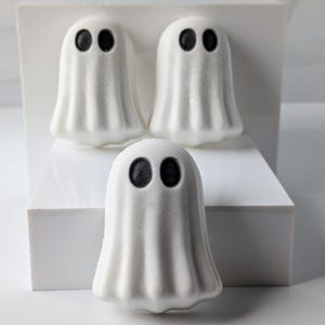 May include: Three white ghost-shaped bath bombs with black eyes are arranged on a white platform. The bath bombs have a textured, slightly sparkly surface, and are designed for Halloween. The background is a minimalist white.