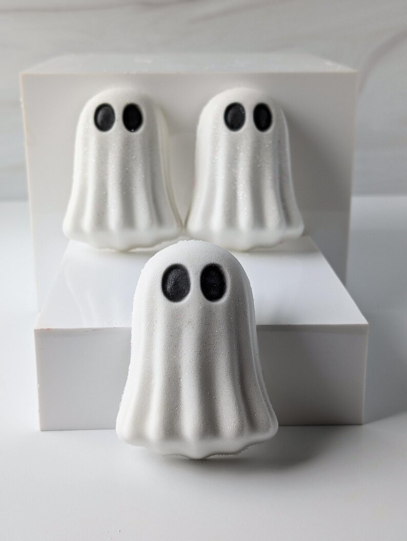 May include: Three white ghost-shaped bath bombs with black eyes are arranged on a white platform. The bath bombs have a textured, slightly sparkly surface, and are designed for Halloween. The background is a minimalist white.