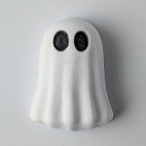 May include: A white, ghost-shaped item with two black, circular eyes. The ghost has a textured, glittery surface. The design is simple and evokes a Halloween theme.