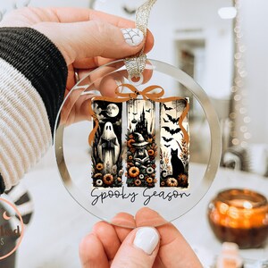 May include: Clear, round Halloween ornament with three vertical panels featuring a ghost, witch, and black cat. The panels are decorated with pumpkins, bats, and a spooky castle. The words "Spooky Season" are written in a script font.