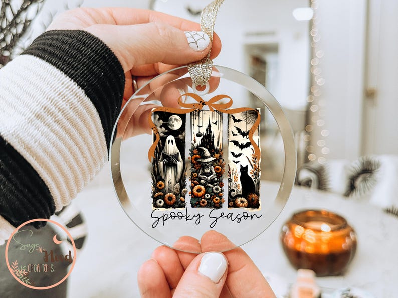 May include: Clear, round Halloween ornament with three vertical panels featuring a ghost, witch, and black cat. The panels are decorated with pumpkins, bats, and a spooky castle. The words "Spooky Season" are written in a script font.