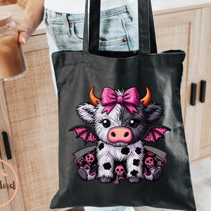 May include: Black tote bag featuring a cartoon cow with a pink bow, horns, and bat wings. The cow has black and white spots and is wearing a dress with skulls. The bag has black handles.
