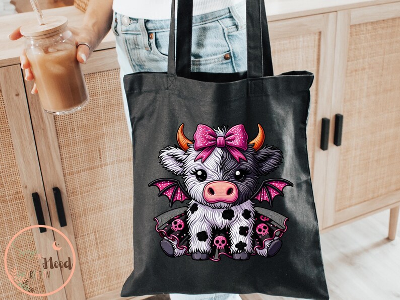 May include: Black tote bag featuring a cartoon cow with a pink bow, horns, and bat wings. The cow has black and white spots and is wearing a dress with skulls. The bag has black handles.
