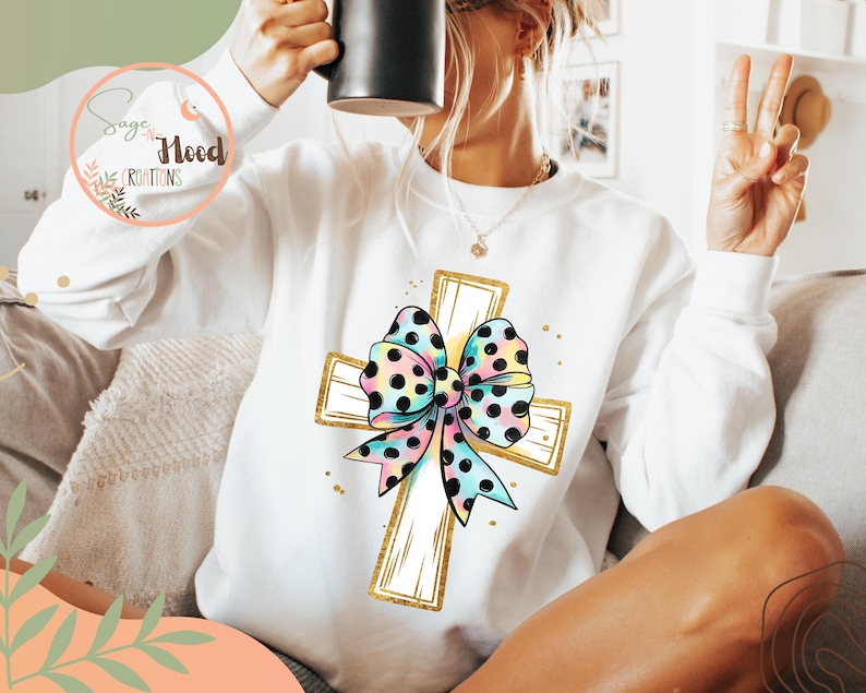 May include: White sweatshirt with a gold cross and a colorful polka dot bow. The bow is tied around the cross.