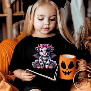 May include: Black t-shirt featuring a cartoon cow with bat wings, a pink bow, and a skull. An orange mug with a jack-o'-lantern face is held in the hand. A large pumpkin is in the background. Halloween theme.