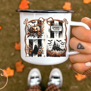 May include: White enamel mug with a black rim and handle. The mug features a Halloween-themed design with a pumpkin, bats, a ghost, and a haunted house. The words "Spooky Season" are also displayed on the mug.