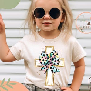 May include: A child wearing a white t-shirt with a gold cross and a colorful polka dot bow. The cross is decorated with gold accents. The child is wearing sunglasses and has their hand raised.