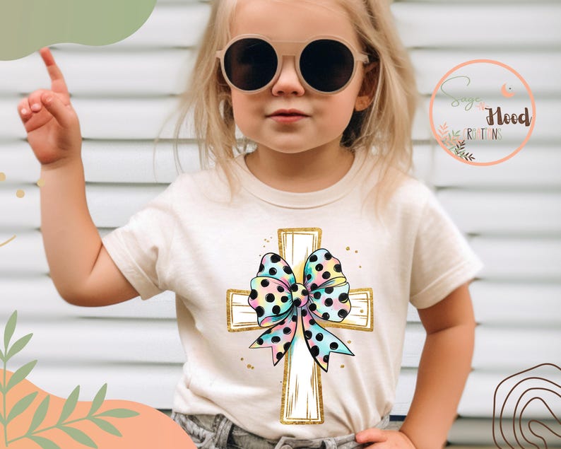 May include: A child wearing a white t-shirt with a gold cross and a colorful polka dot bow. The cross is decorated with gold accents. The child is wearing sunglasses and has their hand raised.
