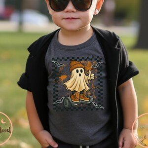 May include: A dark grey t-shirt with a cartoon ghost wearing a beanie and trainers, riding a hoverboard. The ghost gives a thumbs-up. The design includes bats and a checkered background, ideal for Halloween.
