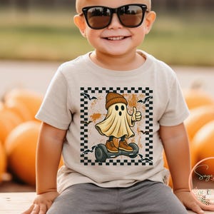 May include: A beige toddler t-shirt featuring a Halloween graphic. The design shows a cartoon ghost wearing a beanie and trainers, riding a hoverboard, giving a thumbs-up. The graphic is framed by a black and white chequered border.