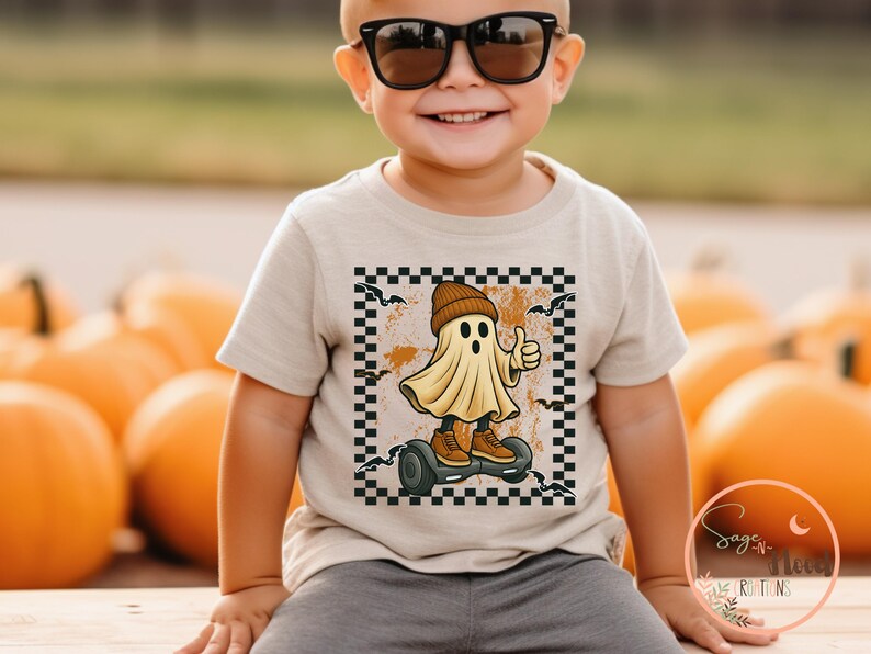 May include: A beige toddler t-shirt featuring a Halloween graphic. The design shows a cartoon ghost wearing a beanie and trainers, riding a hoverboard, giving a thumbs-up. The graphic is framed by a black and white chequered border.