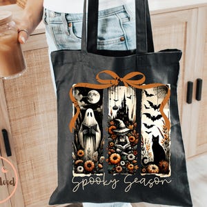 May include: Black tote bag with a Halloween-themed design. The design features a ghost, a witch, a black cat, bats, and a spooky castle. The words "Spooky Season" are written in a script font. The bag is held by a person.