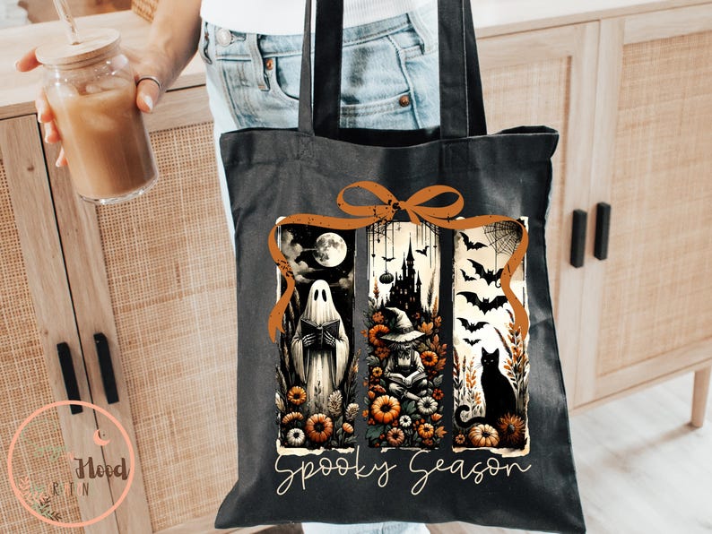 May include: Black tote bag with a Halloween-themed design. The design features a ghost, a witch, a black cat, bats, and a spooky castle. The words "Spooky Season" are written in a script font. The bag is held by a person.